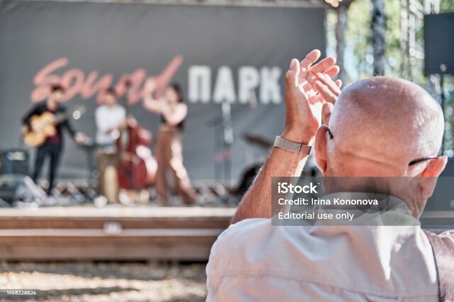 Yekaterinburg, Russia - August 28, 2022: Mature man applauds, raising his hands high. Concert of young jazz group on summer stage in city park. Close-up. Back view. Selective focus